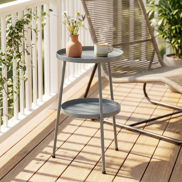 Runder Metall-Beistelltisch mit zwei Ebenen auf einem sonnigen Holzbalkon neben einem Gartenstuhl.