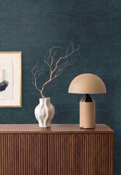 Vliestapete mit Leinenstruktur in Dunkelblau vor einem Sideboard aus Holz mit einer Tischleuchte und einer Vase.