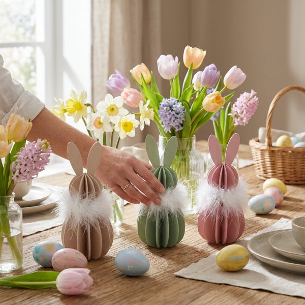 Drei dekorative Osterhasenfiguren aus Holz mit Flauschkragen auf einem Esstisch dekoriert mit Frühlingsblumen und bemalten Ostereiern.