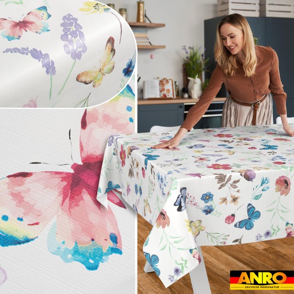 Abwaschbare Wachstuch-Tischdecke mit Schmetterlings- und Blumenmuster auf einem Holztisch in einer hellen Küche. ANRO Deutsche Manufaktur Logo.