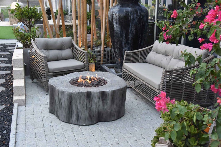 Moderne Gartenlounge mit Feuerschale in Baumstamm-Optik, Polyrattan-Sofa und Sessel auf einer gepflasterten Terrasse.