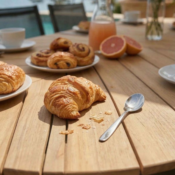 Frühstück auf einem hölzernen Gartentisch mit Croissants, Gebäck und Grapefruit bei hellem Tageslicht.