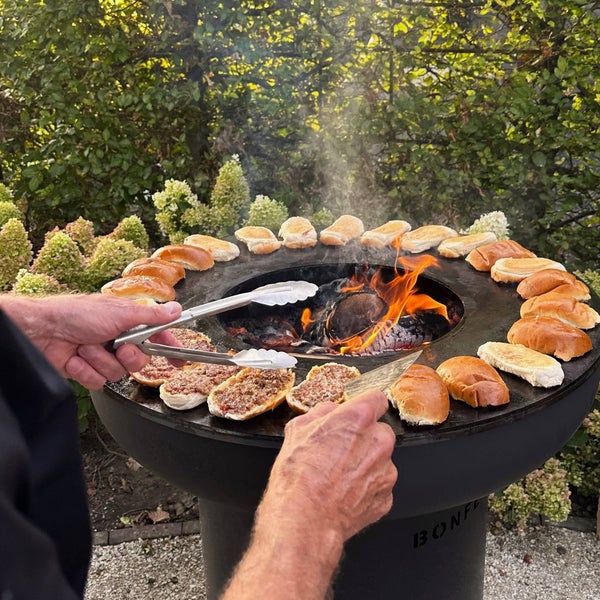 Person grillt Brötchen und Fleisch auf einer runden Feuerschale mit Grillring der Marke BonFeu bei offenem Feuer im Garten.