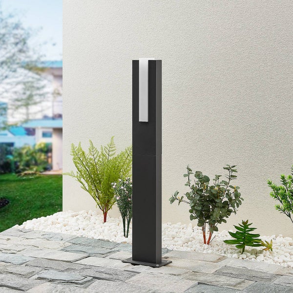 Moderne quadratische Pollerleuchte in einem Gartenbeet mit weißen Ziersteinen und grünen Pflanzen vor einer verputzten Hauswand.