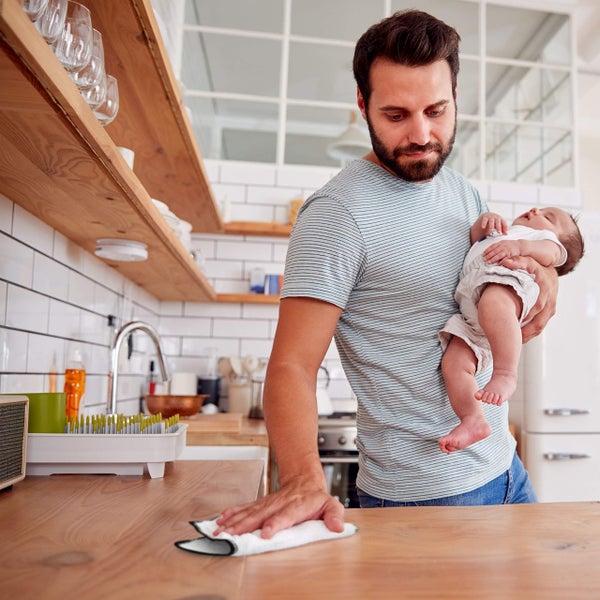 Mann wischt mit einem Tuch eine Arbeitsplatte ab, während er ein Baby hält. Küchenszene.