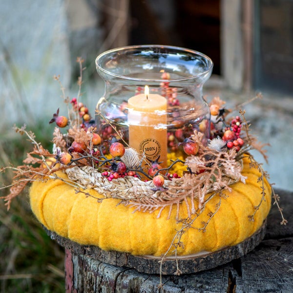 Herbstliche Tischdekoration mit brennender Kerze in Glaswindlicht auf Filzunterlage mit Trockenblumen und Beeren auf einem Baumstumpf.