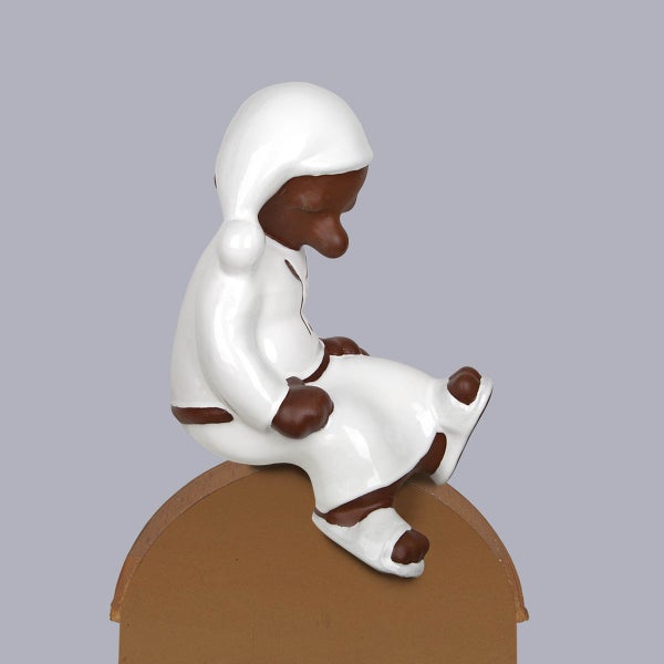 Dekofigur aus Keramik, sitzendes Sandmännchen mit weißer Schlafmütze und Nachthemd auf einem braunen Sockel.