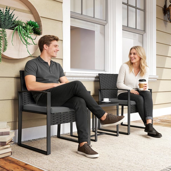 Moderne Outdoor-Sitzgruppe mit zwei schwarzen Polyrattan-Stühlen, grauen Kissen und einem Beistelltisch auf einer hellen Terrasse mit zwei Personen.