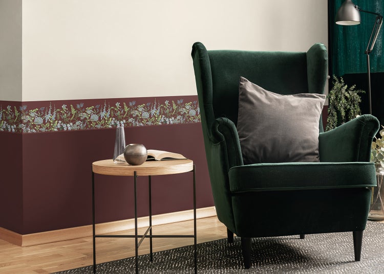 Selbstklebende Tapetenbordüre mit Blumenmuster in einem Wohnzimmer mit grünem Samtsessel und Holzbeistelltisch.
