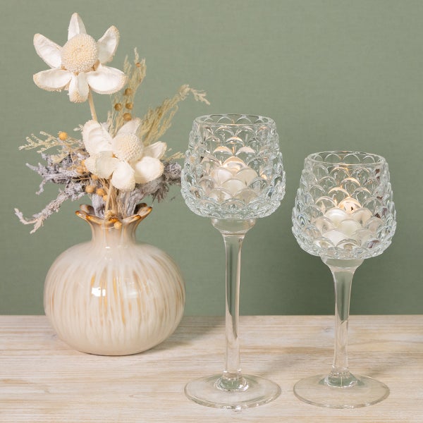 Zwei Teelichthalter aus Glas auf Stielen mit Reliefmuster und eine dekorative Vase mit Trockenblumen auf einem Holztisch.