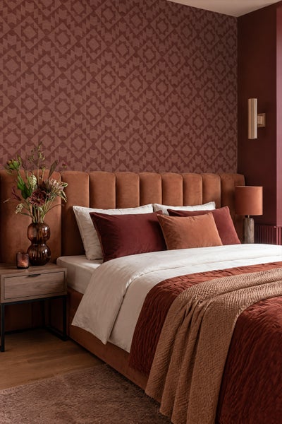 Schlafzimmer mit geometrisch gemusterter Tapete und einem gepolsterten Kopfteil aus Samt in warmen Tönen mit aufeinander abgestimmter Bettwäsche.