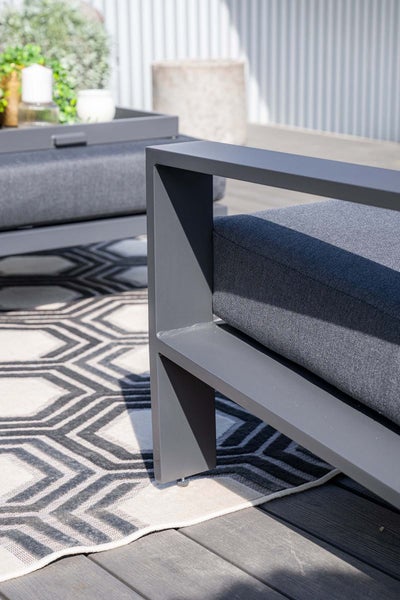 Detailansicht einer modernen Gartenlounge aus Metall mit grauen Polstern auf einem geometrisch gemusterten Teppich.