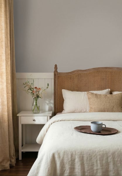 Schlafzimmer mit geflochtenem Kopfteil am Bett, heller Bettwäsche, weißem Nachttisch mit Vase und grauer Wandfarbe über weißer Wandtäfelung.