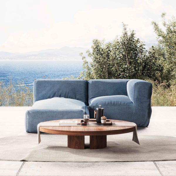Moderne Garten-Lounge mit blauem Sofa und rundem Holztisch auf einer hellen Terrasse mit Meerblick und Bergen im Hintergrund.