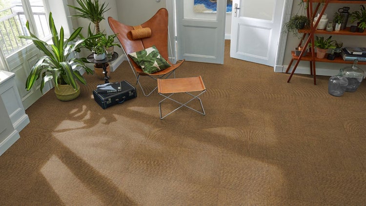 Sisal-Teppichfliesen in einem hellen Wohnzimmer mit Ledersessel und Zimmerpflanzen. Naturfarbene, gewebte Struktur.