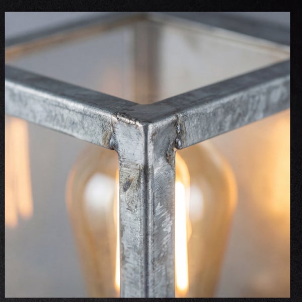 Nahaufnahme einer Lampe im Industriestil mit verzinktem Metallrahmen, Glasfenstern und einem warm leuchtenden Filament-Leuchtmittel.