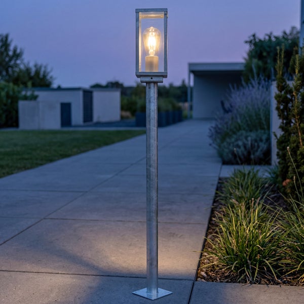 Moderne Außenstandleuchte aus verzinktem Metall mit Glaskopf und leuchtendem Leuchtmittel auf einem Gartenweg in der Abenddämmerung.