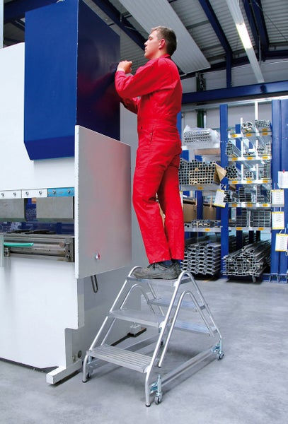 Ein Mitarbeiter in Arbeitskleidung steht auf einer fahrbaren Aluminium Arbeitsplattform vor einer industriellen Biegemaschine in einer Lagerhalle.