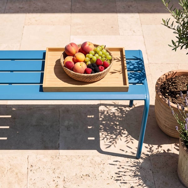Blaue Gartenbank aus Metall mit Holztablett und Obstschale auf einer hellen Terrasse mit Steinboden und Topfpflanzen.