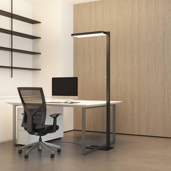 Ein moderner Arbeitsplatz mit einer hohen, schwarzen Stehleuchte, einem weißen Schreibtisch mit Monitor und einem ergonomischen Bürostuhl.
