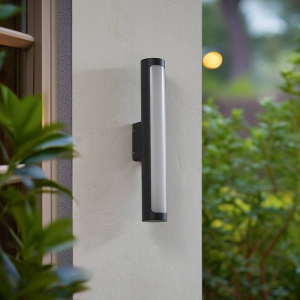 Zylindrische Außenwandleuchte in Schwarz mit vertikalem Diffusor, an einer Hauswand neben einem Fenster montiert.