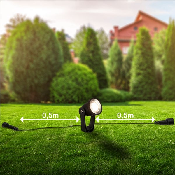 Schwarzer LED-Gartenstrahler auf einem grünen Rasen mit Kabeln von 0,5 Meter auf jeder Seite vor einem Hintergrund mit Bäumen und einem Haus.