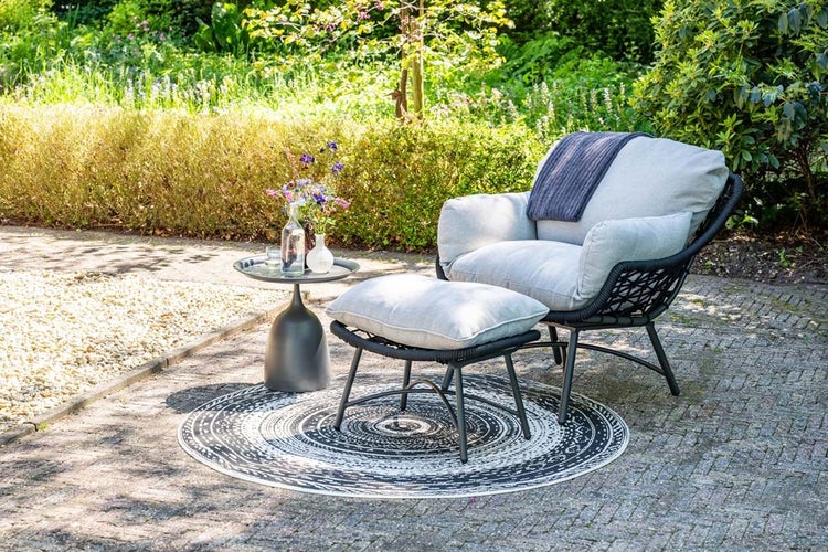 Garten-Lounge-Set auf einer Steinterrasse mit einem geflochtenen Sessel, grauen Polstern, einem Hocker, einem Beistelltisch und einem runden Teppich.