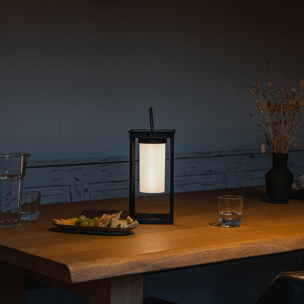 Schwarze LED-Tischleuchte in Laternenform mit warmem Licht auf einem Holztisch in einem gedimmten Raum mit Snacks und Gläsern.