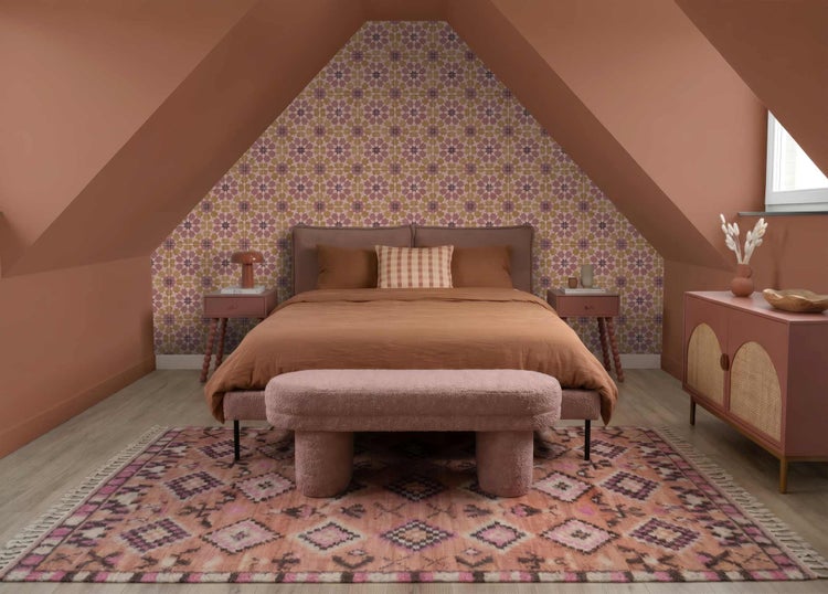 Gemütliches Dachschlafzimmer mit Bett in Terracotta, einer geometrisch gemusterten Tapete, einer rosa Sitzbank und einem gemusterten Teppich.