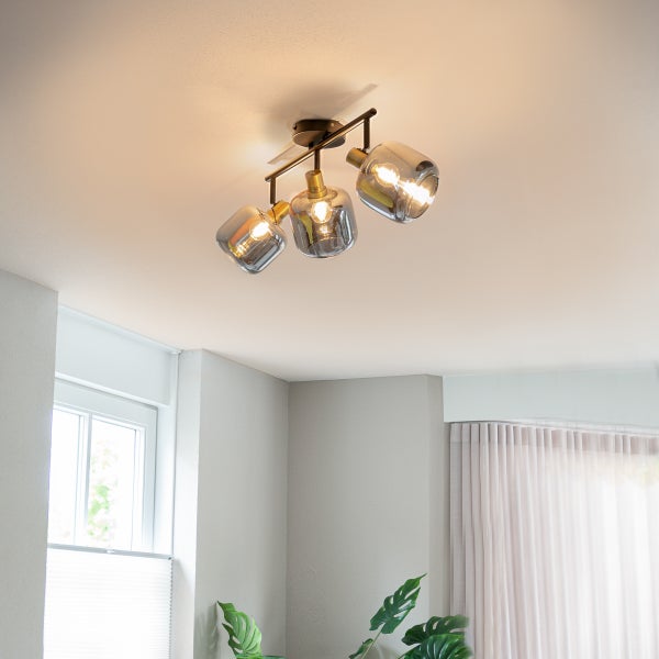 Moderne Deckenleuchte mit drei rauchgrauen Glasschirmen an einem dunklen Metallgestell in einem hellen Zimmer mit Fenster und Zimmerpflanze.
