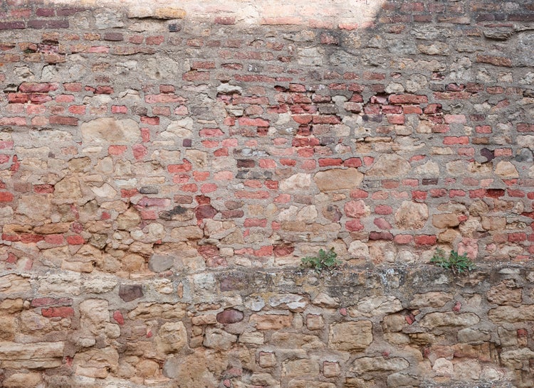 Verwitterte Mauer aus gemischten roten Ziegeln und Natursteinen in verschiedenen Formen und Größen, zusammengehalten durch groben Mörtel.