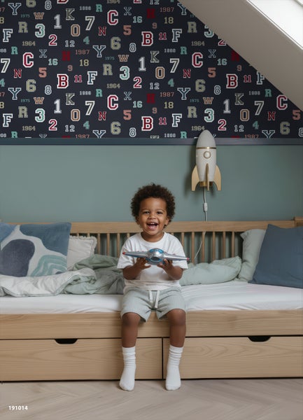 Kinderzimmer mit dunkelblauer Tapete mit bunten Buchstaben und Zahlen. Ein Kind sitzt auf einem Holzbett. Raketenlampe an der Wand.