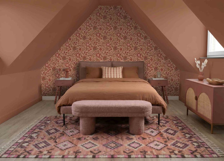 Ein gemütliches Schlafzimmer unter Dachschrägen mit terracottafarbenen Wänden, floraler Tapete, einem gepolsterten Bett und gemustertem Teppich.