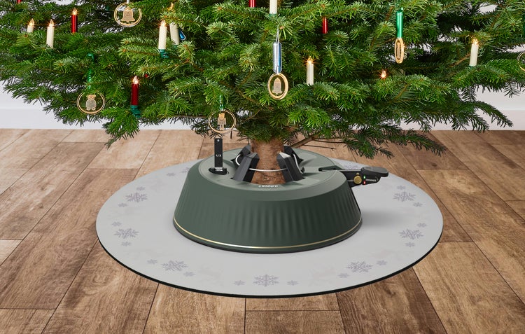Grüner Krinner Christbaumständer mit Fußpedal und Wasserstandsanzeige auf einer grauen Matte unter einem geschmückten Baum auf Holzfußboden.