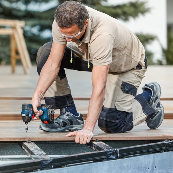 Ein Handwerker kniet und verschraubt eine Holzterrasse mit einem Akku-Schlagschrauber.