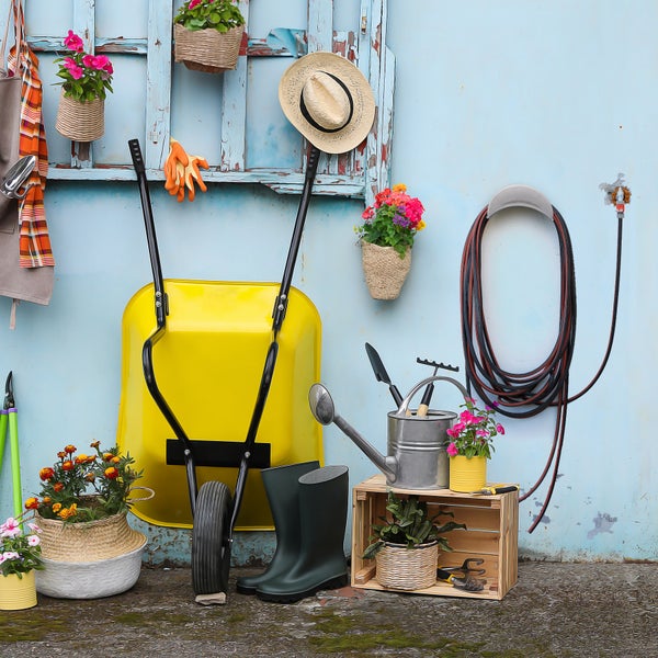 Gartenszene mit Schubkarre, Gießkanne, Gartengeräten, Pflanzen und Accessoires vor einer blauen Wand