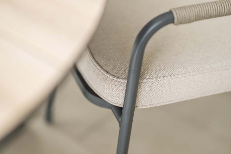 Nahaufnahme eines Gartenstuhls mit gepolsterter Sitzfläche, Metallrahmen und seilumwickelter Armlehne neben einem Holztisch.