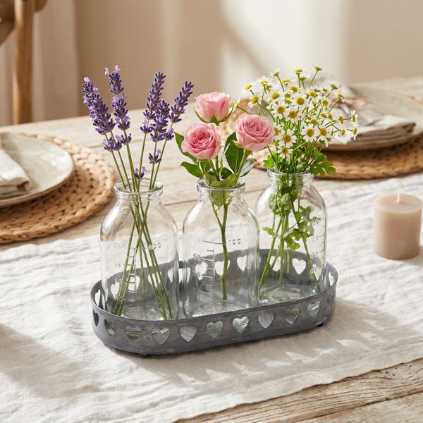 Drei Glasflaschen als Vasen in einem ovalen Metallhalter mit Herzmotiven auf einem Leinentischläufer, dekoriert mit Lavendel, Rosen und Kamille.