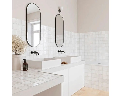 Modernes Badezimmer mit zwei weißen Aufsatzwaschbecken, ovalen Spiegeln mit schwarzem Rahmen, schwarzen Wandarmaturen und weißen quadratischen Fliesen.
