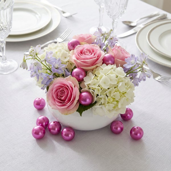 Blumengesteck in einer runden Schale mit Rosen, Hortensien und kleinen Dekokugeln auf einem gedeckten Tisch.