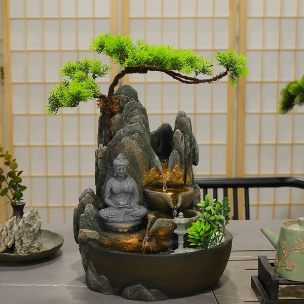 Zimmerbrunnen in Steinoptik mit sitzender Buddha-Figur, künstlichem Bonsai-Baum und fließendem Wasser in beleuchteten Kaskadenbecken.