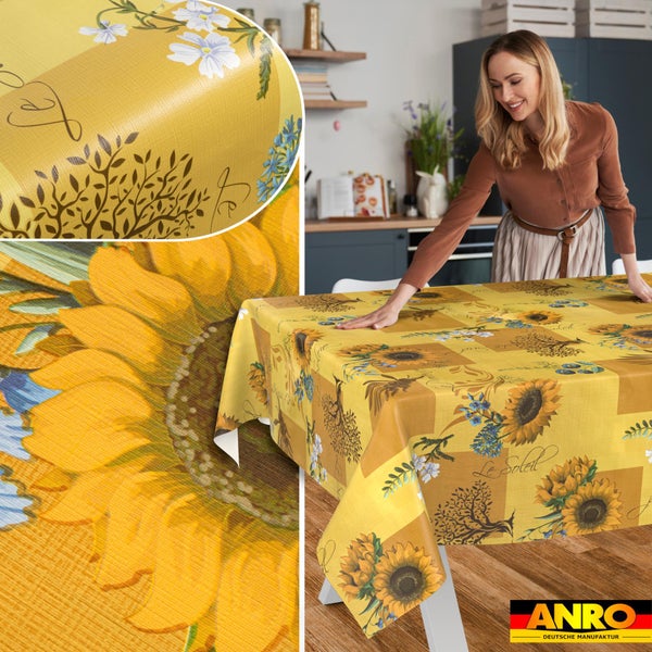 Frau breitet gelbe Wachstuch-Tischdecke mit Sonnenblumen- und Baummuster auf einem Tisch in der Küche aus. Detailansicht Textur. ANRO Logo.