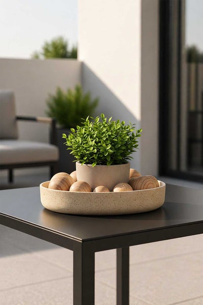 Grüne Pflanze im Topf mit dekorativen Holzkugeln in einer runden Schale auf einem schwarzen Metalltisch auf einer hellen Terrasse.