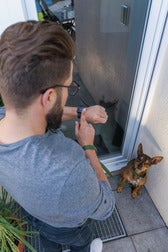 Ein Mann mit Bart und Brille schaut vor einer Tür auf seine Smartwatch, während er einen kleinen Hund an der Leine hält.
