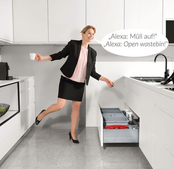 Frau in moderner weißer Küche bedient ein Abfalltrennsystem per Sprachbefehl, wobei sich die Schublade automatisch öffnet.