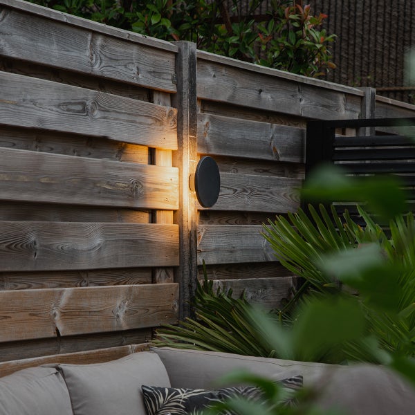 Moderne, runde Außenwandleuchte an einem Holzzaun mit warmem Lichtschein in einem Garten mit Pflanzen und Sitzbereich.