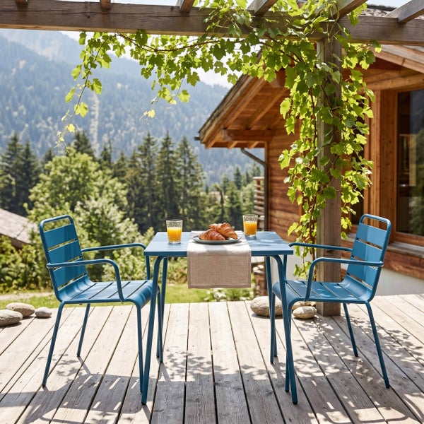 Gartenmöbel-Set aus blauem Metall mit einem quadratischen Tisch und zwei Stühlen auf einer Holzterrasse mit Bergblick.