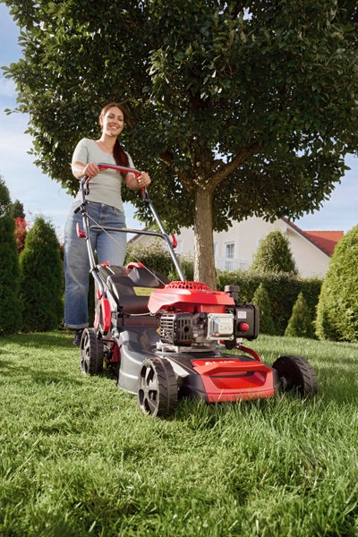 Eine Frau mäht den Rasen mit einem rot-schwarzen Benzin-Rasenmäher in einem hellen Garten mit einem Baum und Sträuchern.