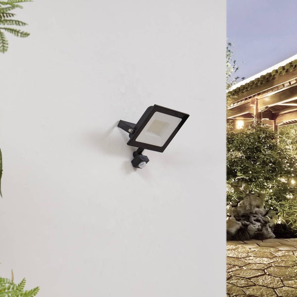 Schwarzer LED-Außenstrahler mit Bewegungsmelder an einer Wand montiert, im Hintergrund ein beleuchteter Garten mit Steinweg und Pflanzen.