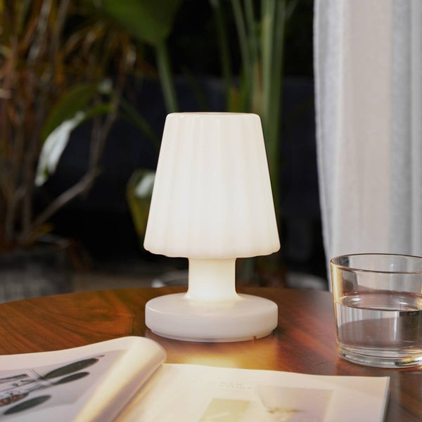 Eine weiß leuchtende LED-Tischleuchte mit plissiertem Schirm auf einem Holztisch neben einem Wasserglas und einem Magazin vor Pflanzen.